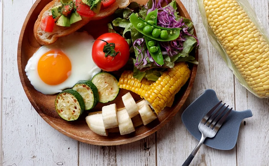 ワンプレートのディッシュに、サンド、トマト、目玉焼き、バナナ、とうもろこしなど、さまざまなカラフルで色彩豊かな野菜を取り入れた健康的な食事のイメージ画像