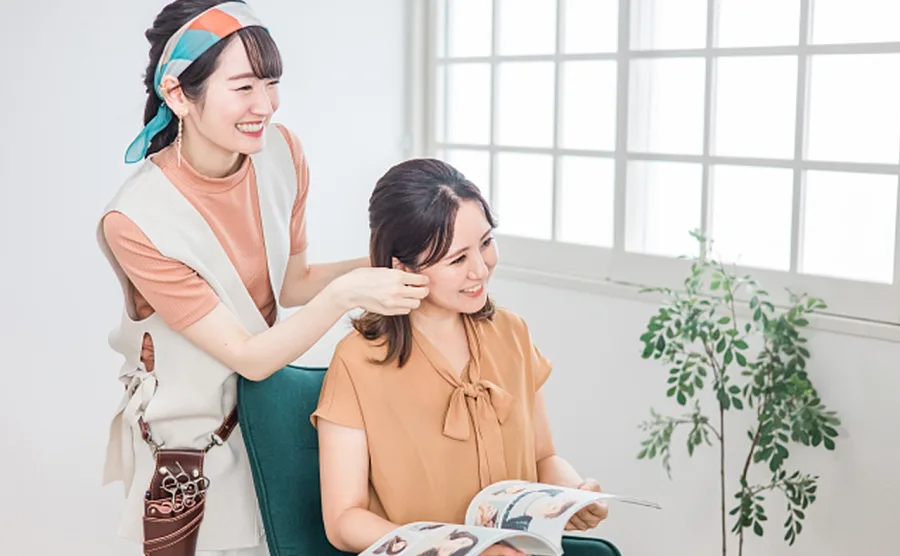 通い慣れた美容室で若い美容師と手に雑誌を持ち、笑顔で話している40代の女性の画像